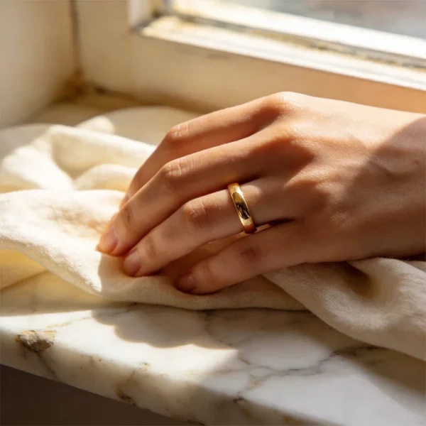 A hand wearing an 18K yellow gold band ring resting on marble, showing the warm rich colour of high karat gold jewellery